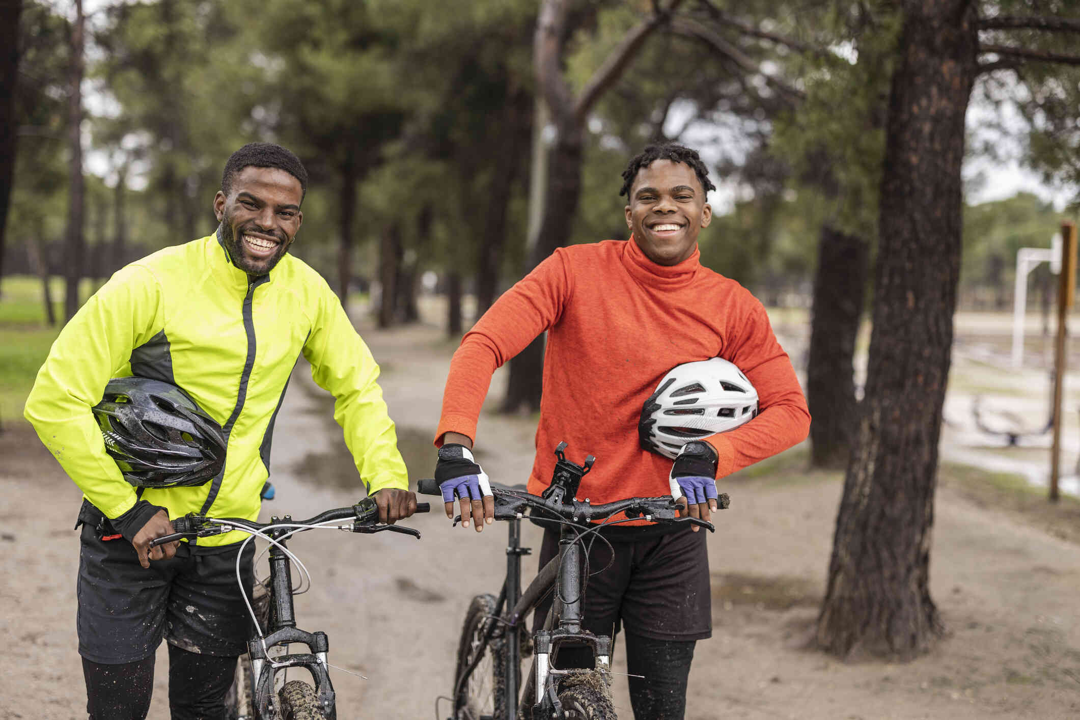 Two adult male friends stand next to each other outside with their bikes while smiling at the camera.